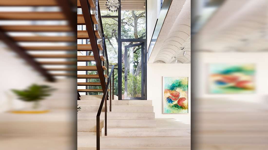 Modern interior entryway with floating wooden staircase featuring black metal railing, large floor-to-ceiling glass door framed in black, and white walls adorned with colorful abstract artwork.
