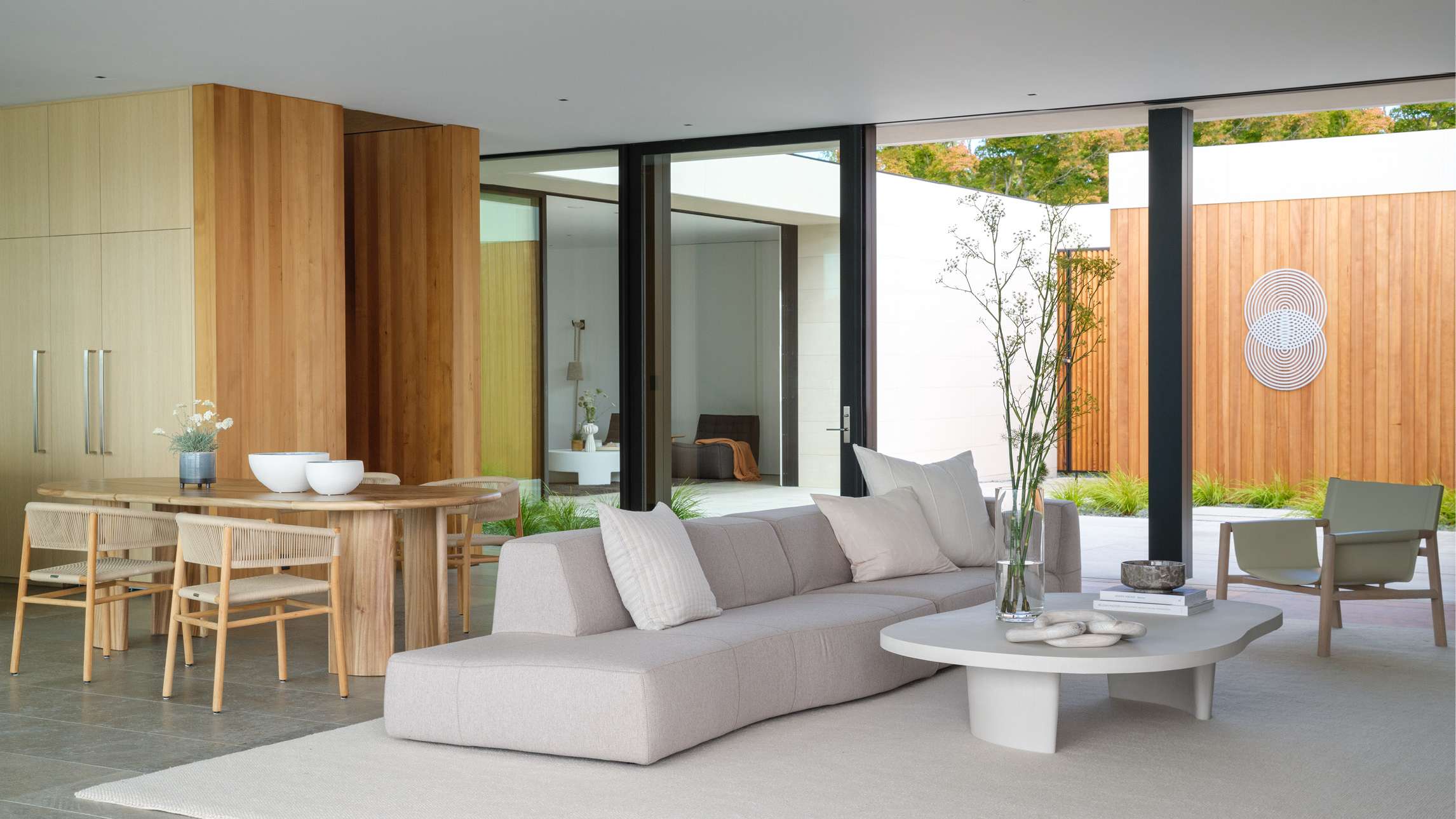 Luxury open-concept living and dining area with light wood paneling, floor-to-ceiling glass doors opening to a courtyard, minimalist beige sectional sofa, sculptural white coffee table, and natural wood dining set.