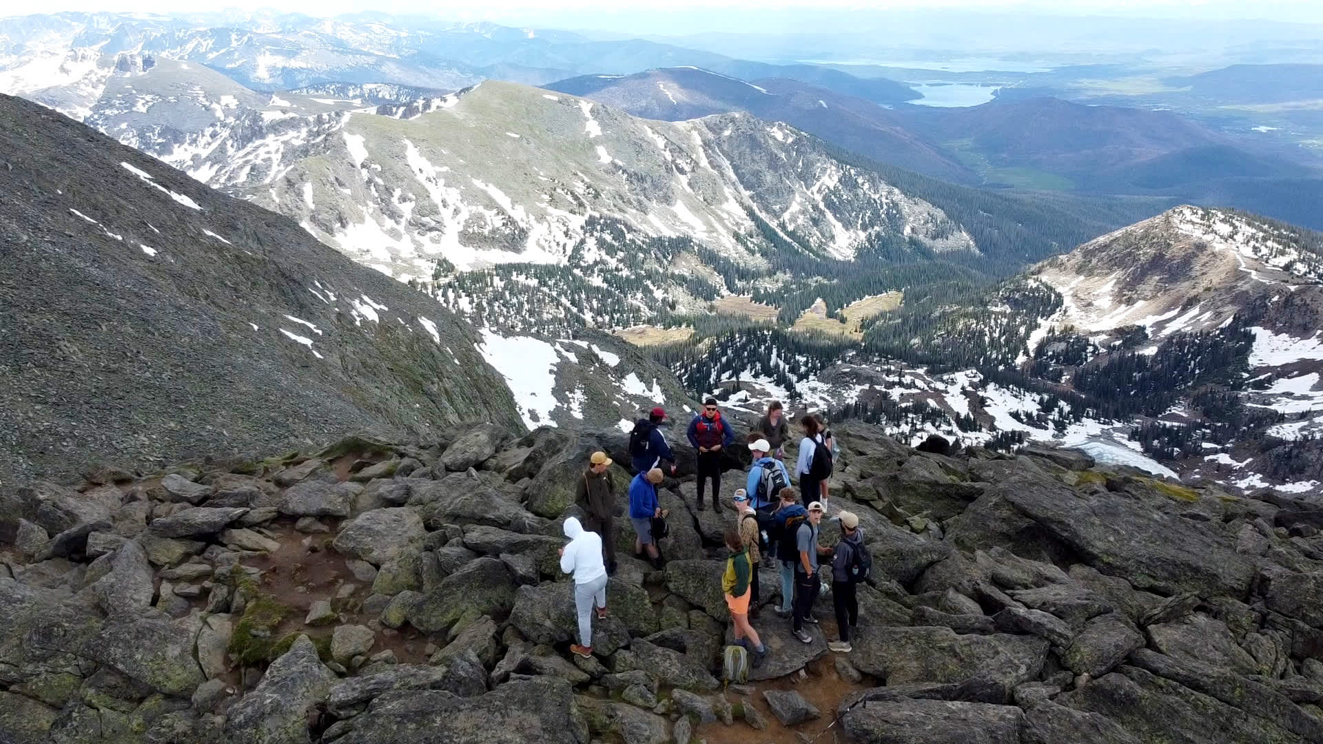 Group of hikers standing on rocky terrain near a mountain summit with snow patches, overlooking a panoramic view of rugged peaks, forested valleys, and distant lakes under a clear sky.