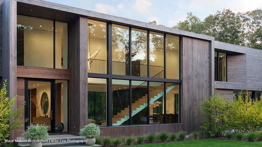 Contemporary two-story home with floor-to-ceiling glass windows, dark wood siding, and open staircase visible through expansive glass facade, surrounded by landscaped greenery.