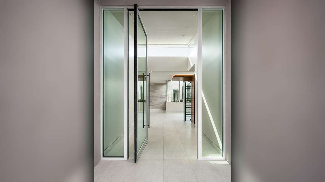 Minimalist entryway with pivot glass door framed by frosted glass panels, light gray tile flooring, and open interior featuring natural light and modern decor elements.