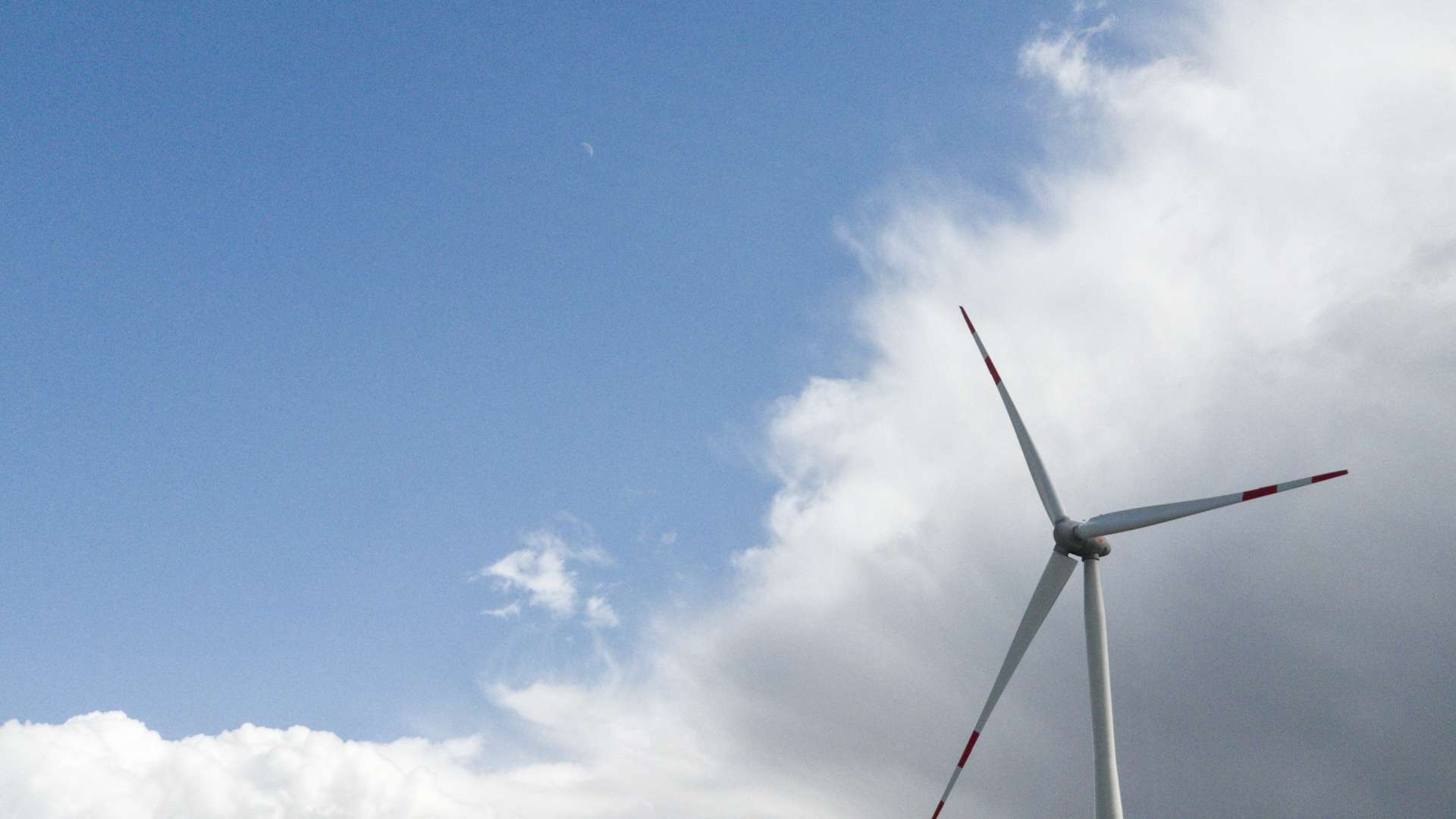 Wind turbine with red tips standing against a bright blue sky and large white clouds in the background.
