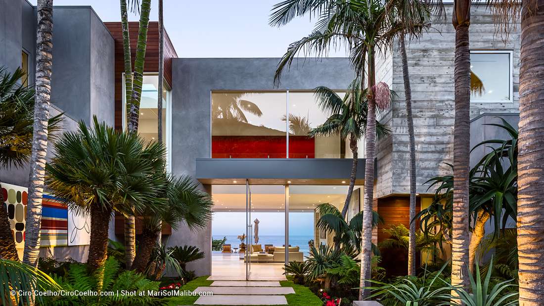 Luxury modern coastal home exterior with minimalist concrete walls, glass balcony railings, vertical wooden slats, and floor-to-ceiling glass doors framed by tropical landscaping featuring palm trees and ocean views.