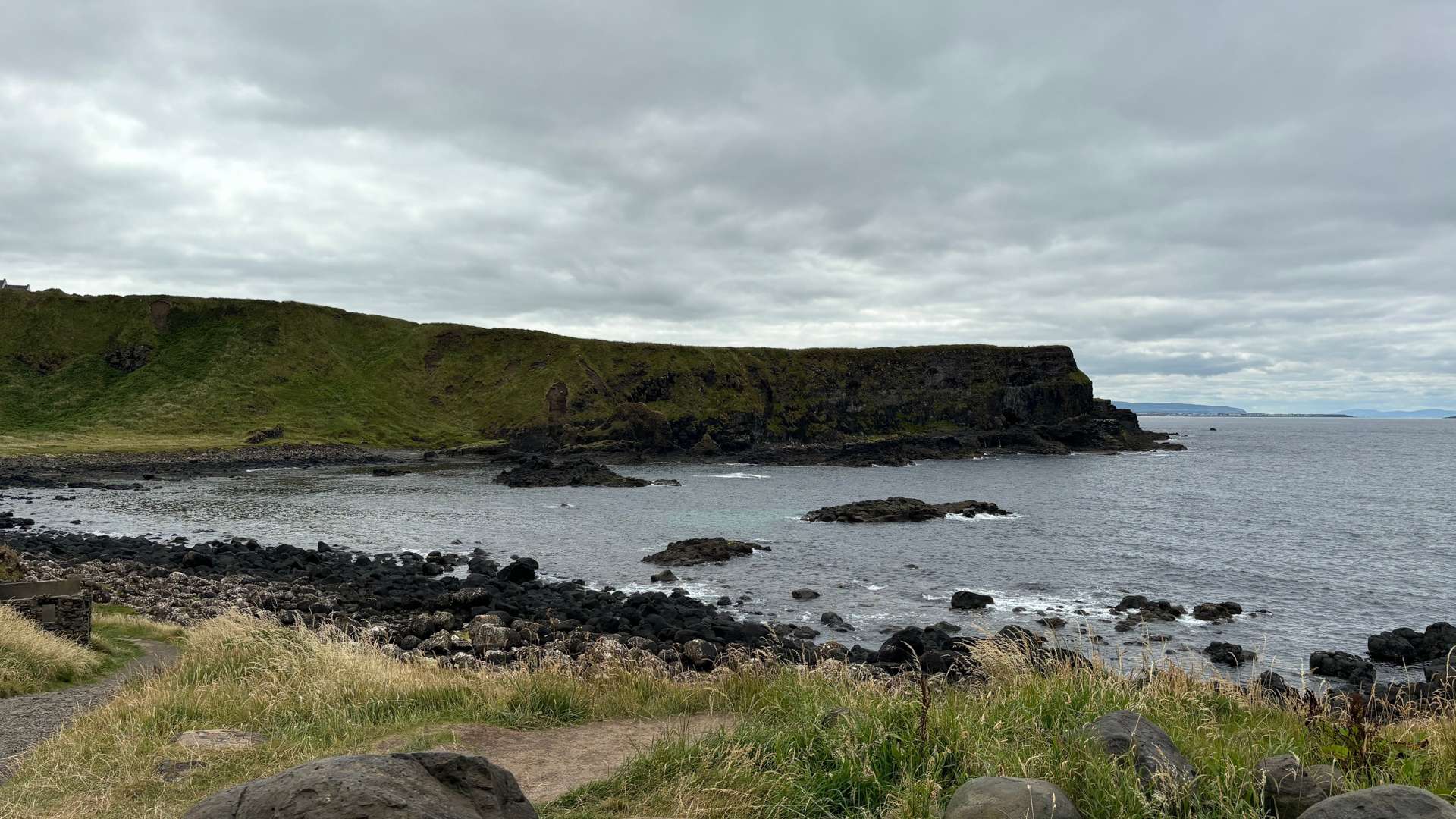 Rocky coastline with grassy cliffs and scattered boulders along calm sea under an overcast sky.