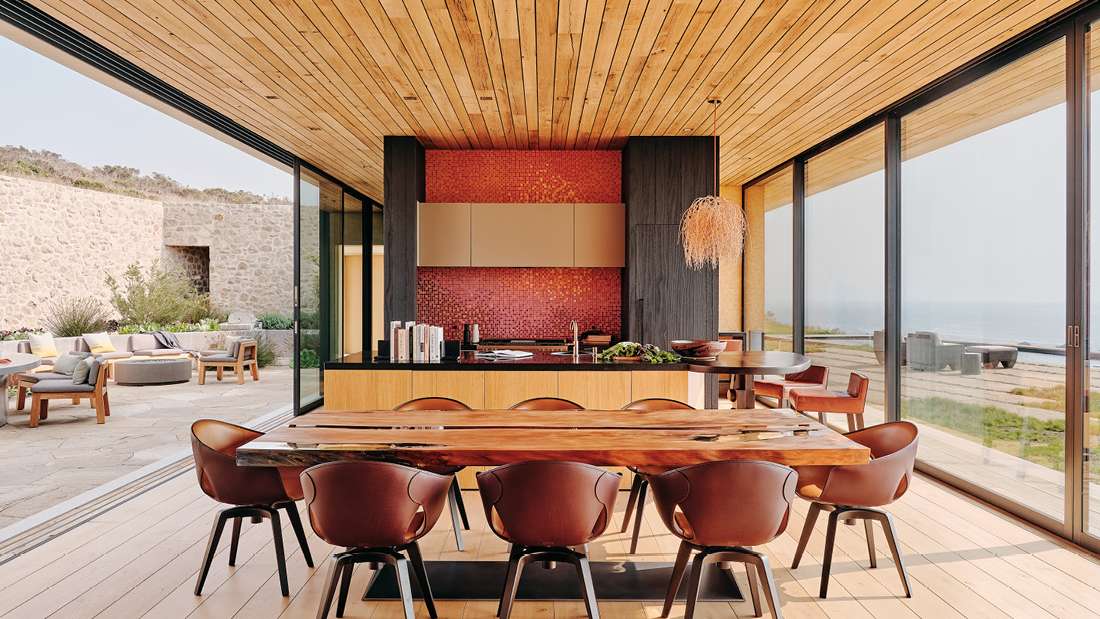 Luxury open-concept kitchen and dining area with natural wood ceiling, large glass sliding doors opening to ocean-view patio, red mosaic tile backsplash, black countertop island, and rustic wooden dining table with leather chairs.