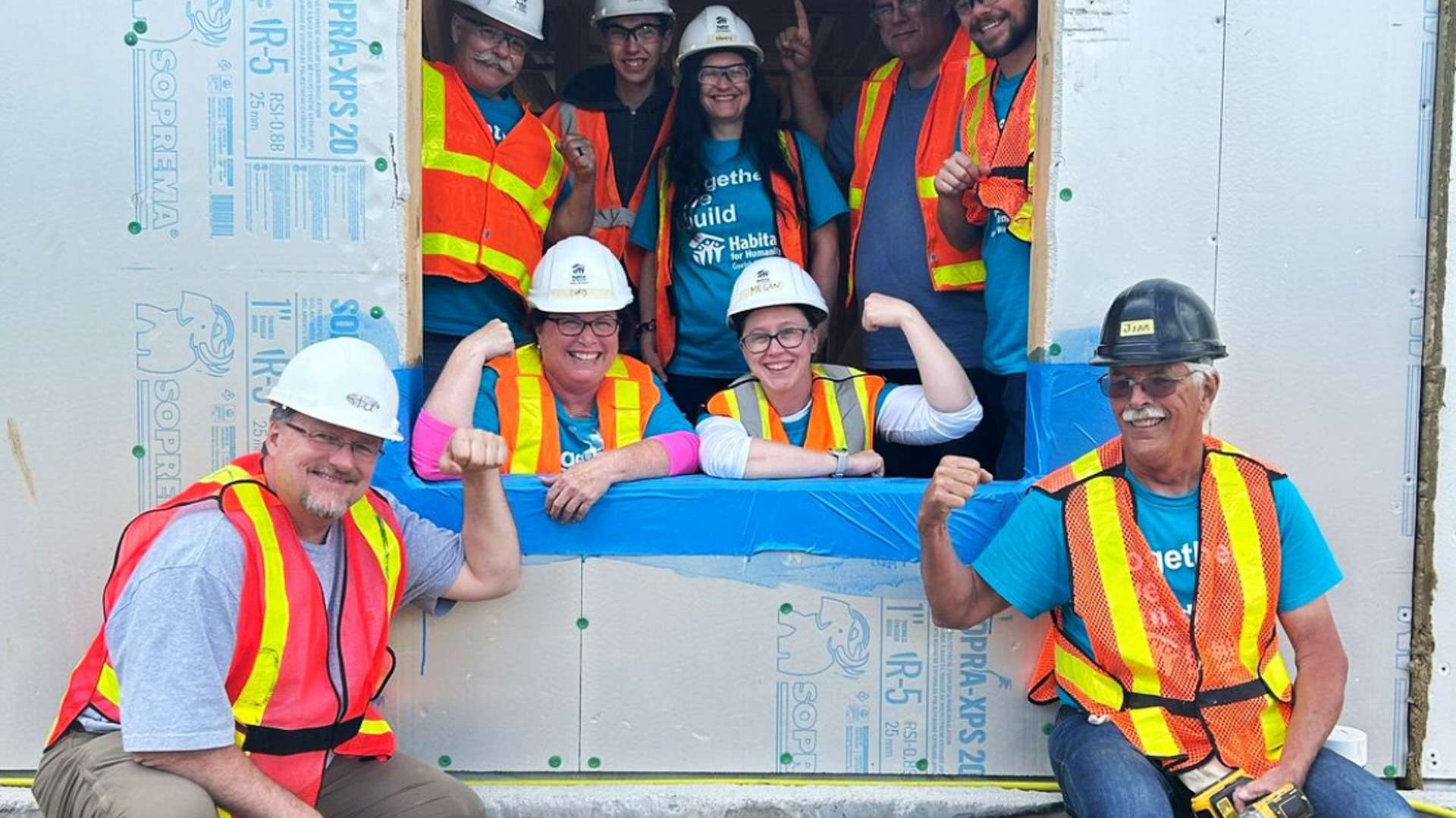 Group of volunteers wearing safety vests and hard hats posing around a window opening of a partially built structure, with some flexing arms and others holding tools.