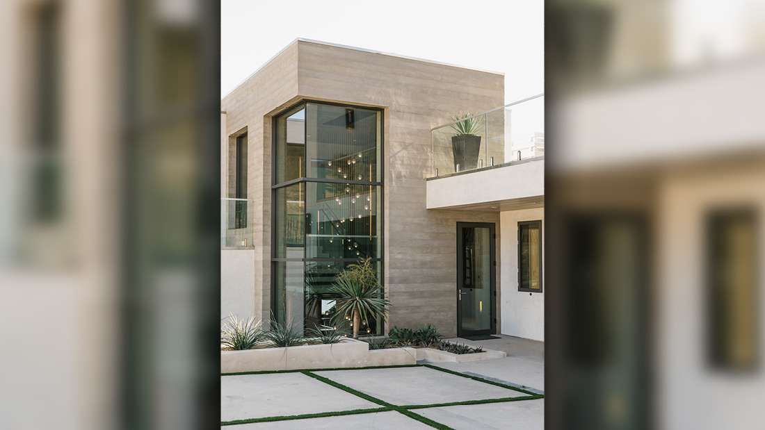 Contemporary two-story home with smooth concrete exterior, floor-to-ceiling glass windows, glass balcony railing, black-framed entry door, and minimalist landscaping featuring desert plants.