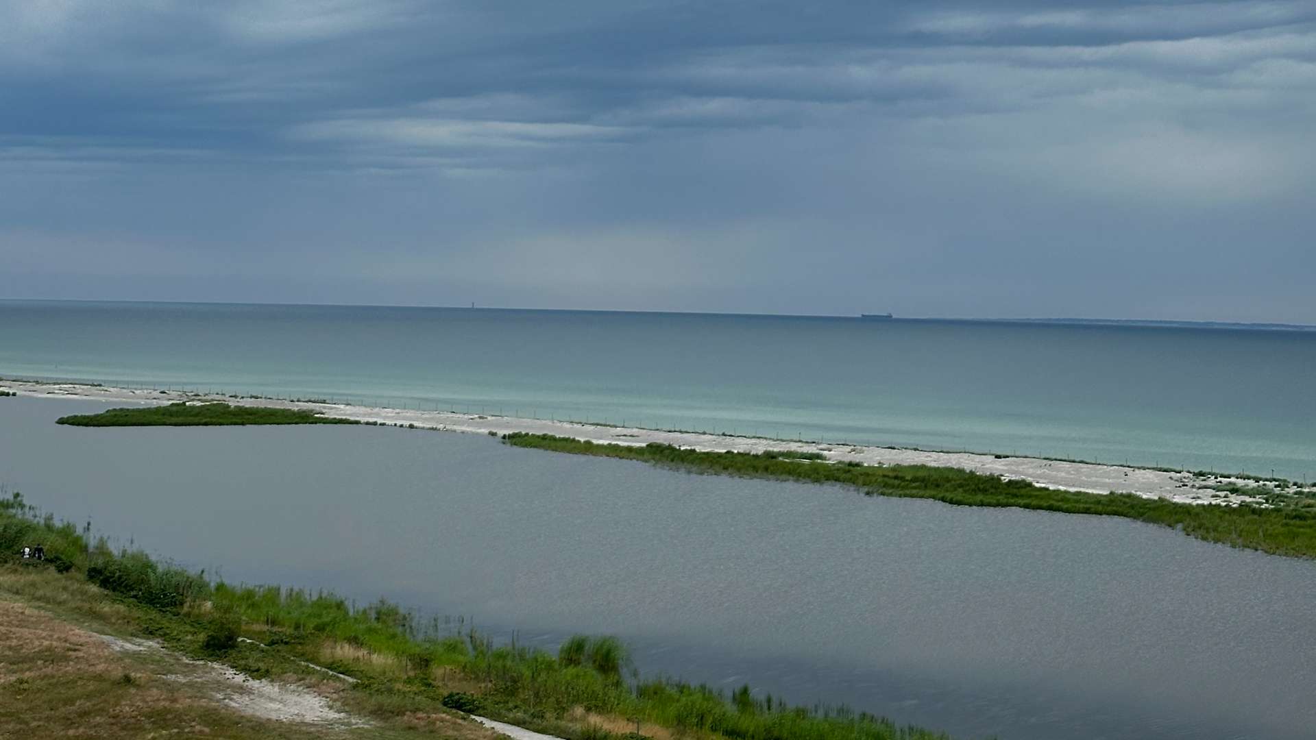 Coastal lagoon with grassy shoreline and sandy beach leading to calm turquoise sea under cloudy skies.