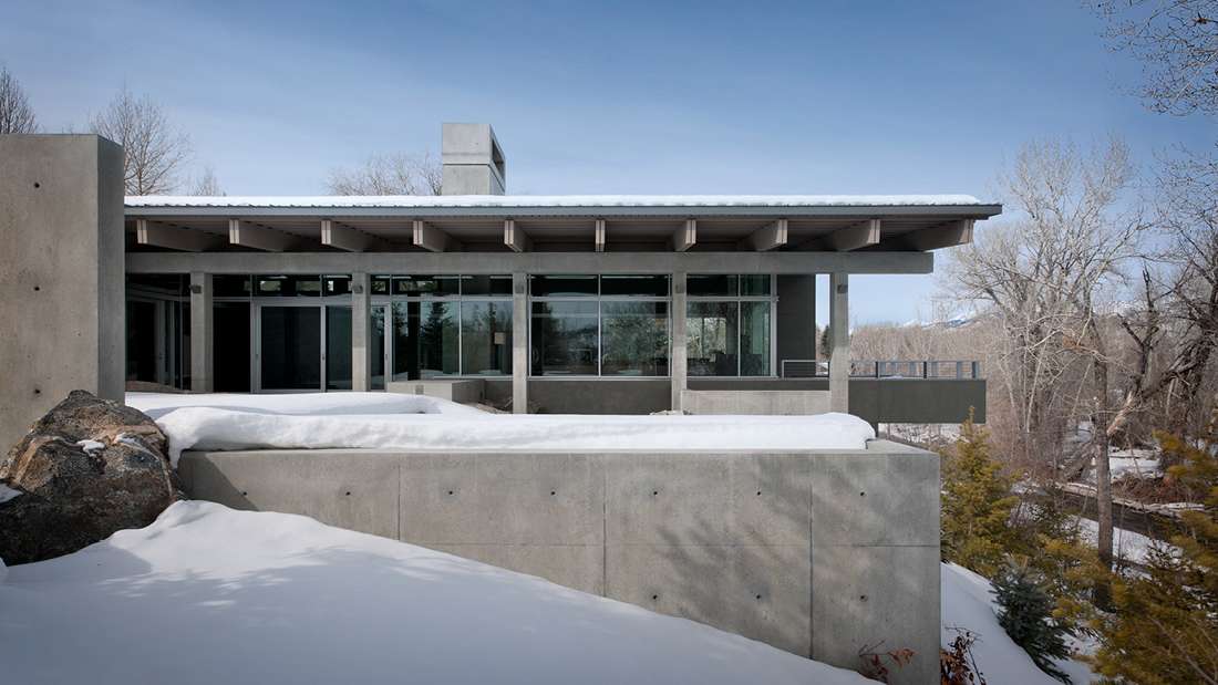 Modern mountain home with concrete exterior walls, flat roof design, and large glass windows surrounded by snow-covered landscape and bare winter trees.