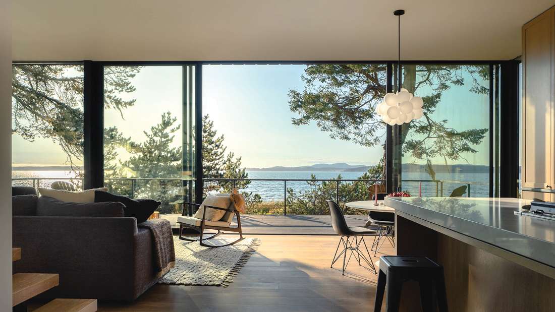 Modern open-concept living and dining area with floor-to-ceiling glass walls, panoramic ocean views, minimalist gray sofa, round dining table with black chairs, and sculptural white pendant light.
