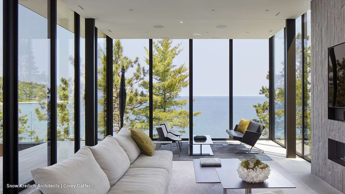 Luxury modern living room with floor-to-ceiling glass walls offering panoramic lake views, light gray sectional sofa, black leather lounge chair, minimalist coffee tables, and stone accent wall with mounted TV.