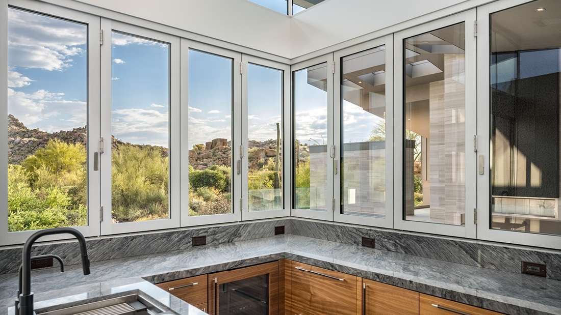 Modern kitchen corner with panoramic desert views through floor-to-ceiling glass windows, gray marble countertops, black faucet, and light wood cabinetry.