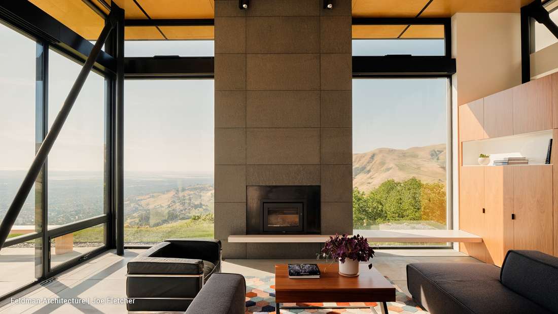 Modern living room with floor-to-ceiling glass walls offering panoramic mountain and valley views, minimalist concrete fireplace, black leather armchair, gray sectional sofa, and wooden cabinetry.