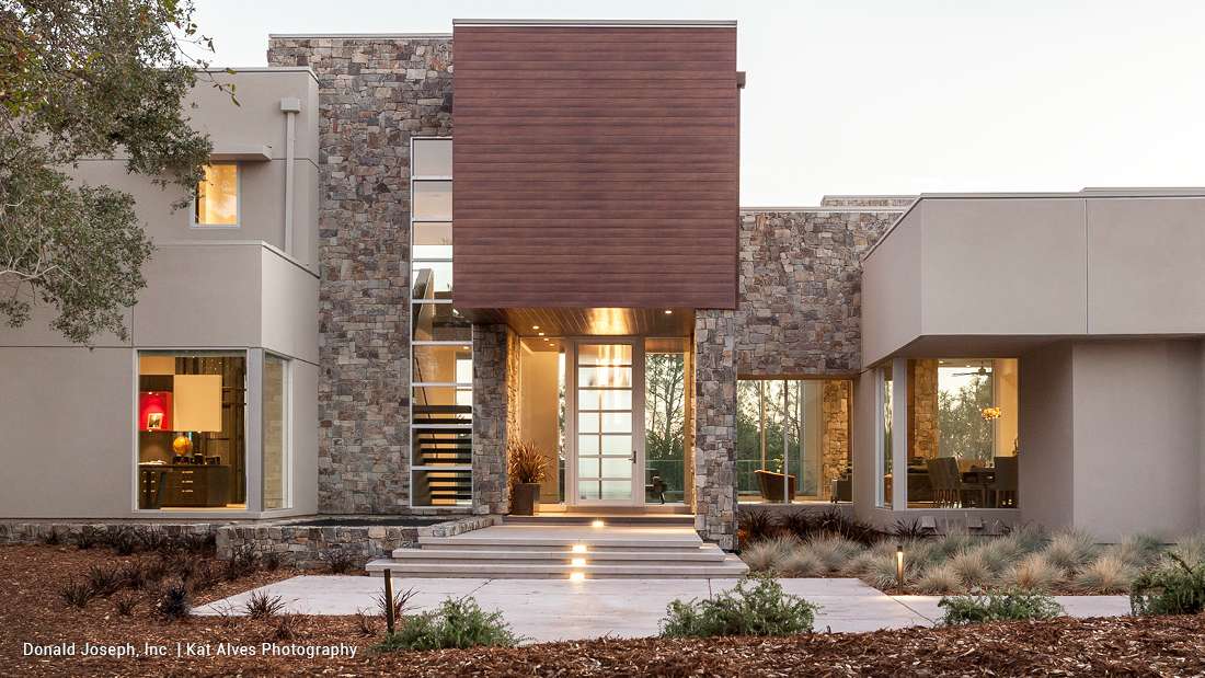 Modern luxury home exterior with stone cladding, large glass windows, and central wooden accent panel above glass entry door, featuring symmetrical design and landscaped front yard.
