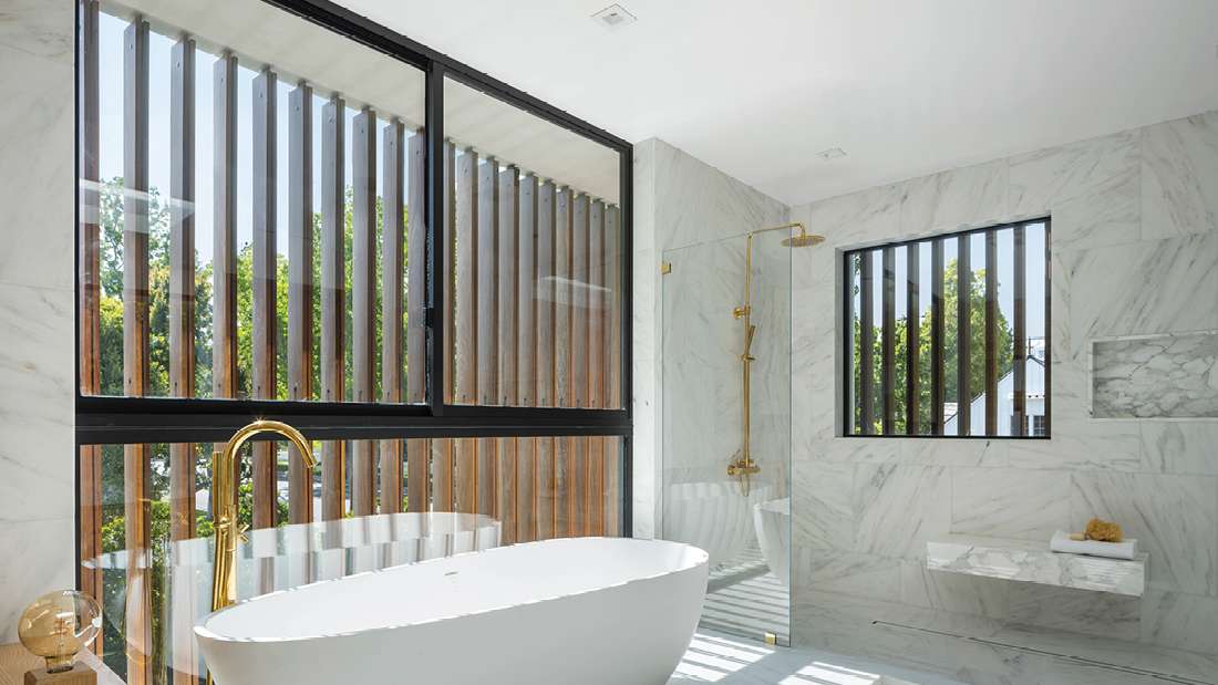 Luxury bathroom with freestanding white oval bathtub, marble walls, large black-framed windows with vertical wooden slats, and glass-enclosed shower featuring gold fixtures.