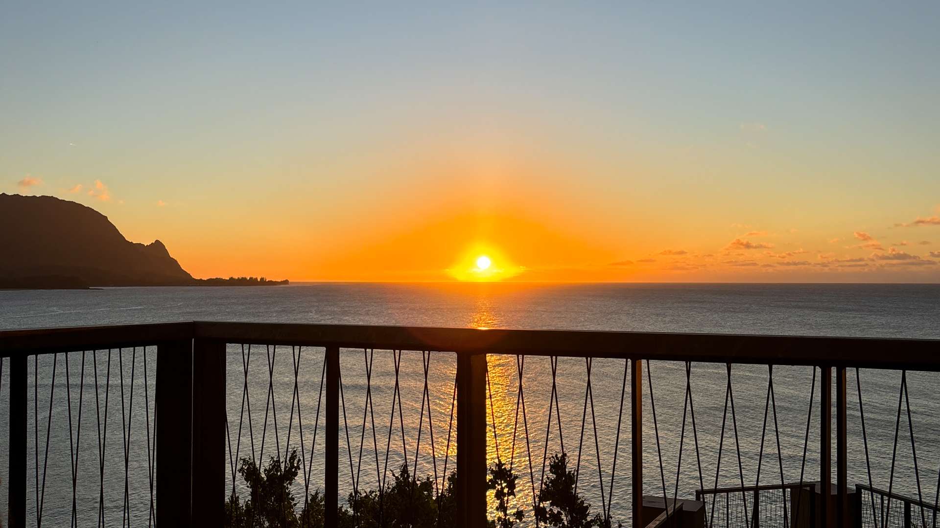 Golden sunset over calm ocean viewed from a balcony with railing and distant mountains on the horizon.