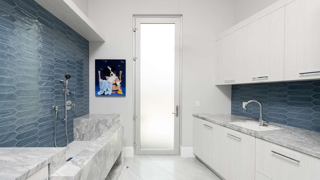 Modern laundry and pet wash room with blue textured tile backsplash, marble countertops, built-in white cabinetry, and frosted glass door providing natural light.