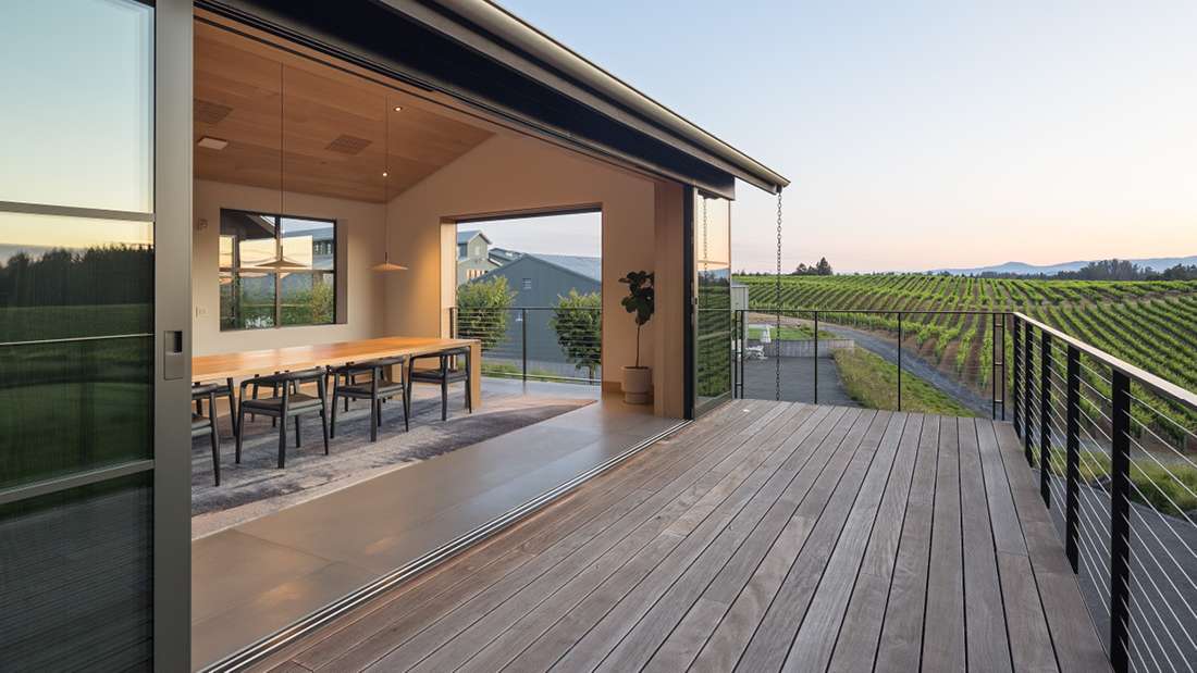 Modern dining area with long wooden table and black chairs, sliding glass doors opening to a spacious deck overlooking vineyard rows and distant hills at sunset.