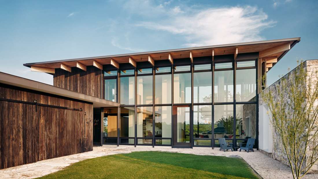 Contemporary home exterior with dark wood siding, expansive floor-to-ceiling glass walls, sloped roof with exposed beams, and landscaped yard featuring green lawn and gravel walkway.