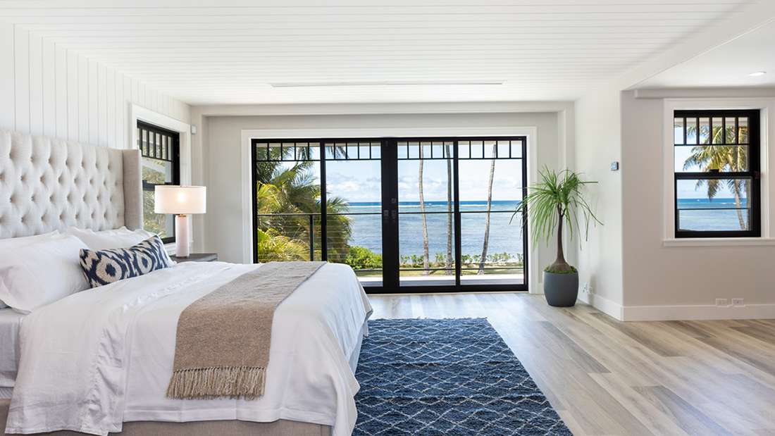 Coastal bedroom with tufted headboard, white bedding, blue patterned rug, and sliding glass doors opening to ocean views and palm trees.