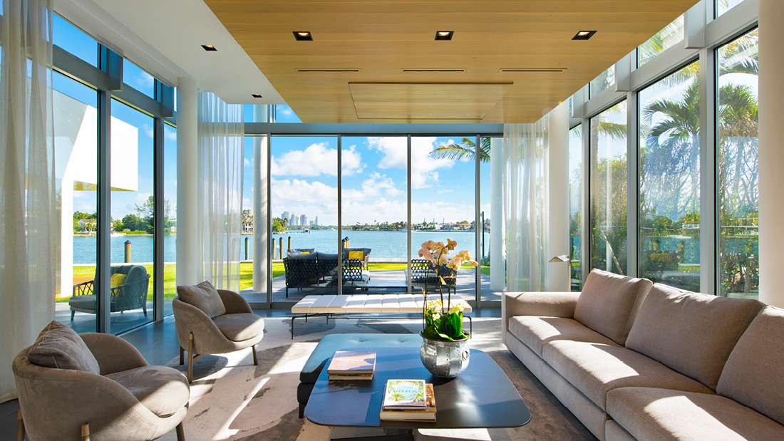 Luxury waterfront living room with floor-to-ceiling glass walls offering panoramic bay views, light wood ceiling with recessed lighting, sheer white curtains, and modern furniture including beige sofa and curved lounge chairs.