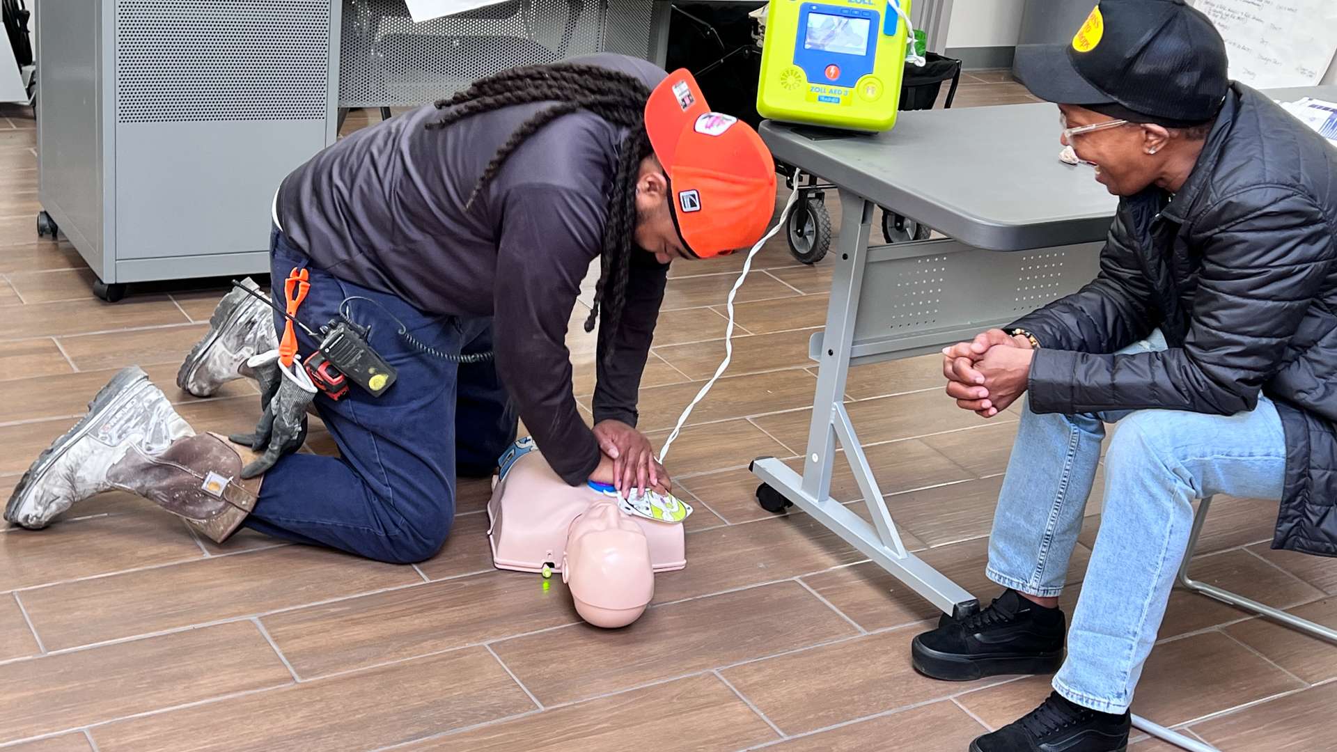 Person kneeling on a wood-look floor performing chest compressions on a CPR manikin during a training session, with an AED device on a nearby table and another participant observing.
