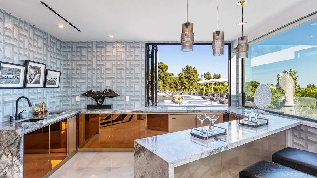 Luxury home bar with marble countertops, mirrored cabinets, geometric patterned wallpaper, three metallic pendant lights, and folding glass window opening to outdoor patio with greenery.