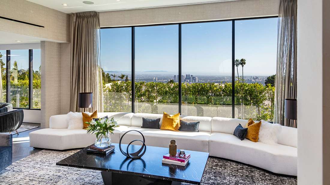 Luxury living room with floor-to-ceiling glass walls offering panoramic city views, long white sectional sofa with colorful accent pillows, black marble coffee table with decorative books and sculpture, and neutral textured curtains.