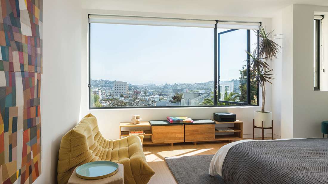 Modern bedroom with large picture window offering panoramic city views, mustard yellow tufted lounge chair, colorful geometric wall art, wooden storage bench with cushions, and potted indoor plant.