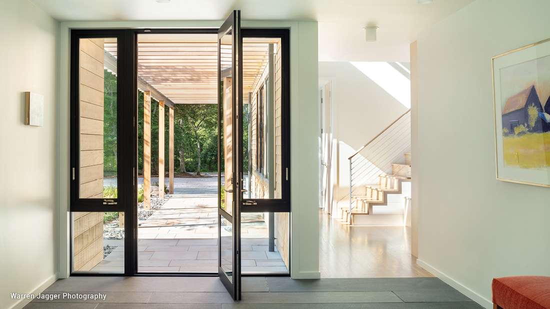 Modern entryway with black-framed glass pivot door opening to a covered walkway featuring horizontal wood slats, light gray tile flooring, and interior staircase with cable railing illuminated by natural light.