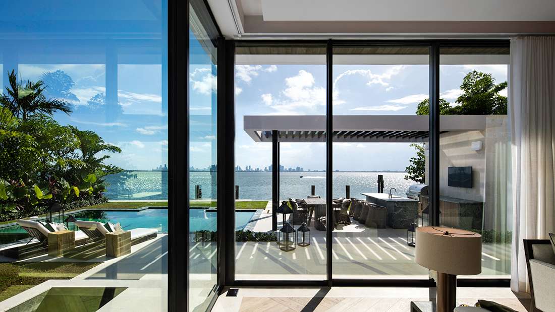 Luxury waterfront home interior with floor-to-ceiling sliding glass doors opening to a patio featuring outdoor dining under a modern pergola, infinity-edge pool, and panoramic view of the skyline.