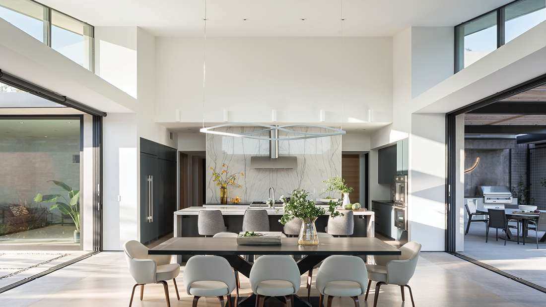 Luxury modern kitchen and dining area with marble backsplash, large island featuring integrated sink, contemporary pendant light fixture, and sliding glass doors opening to outdoor patio with built-in grill.