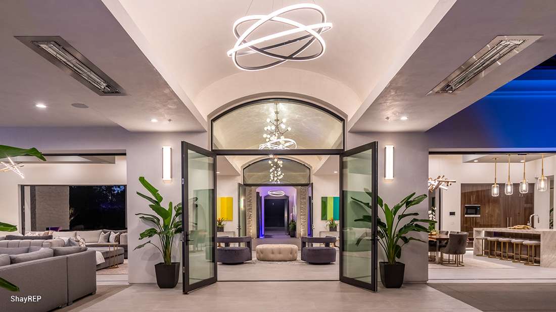 Luxury modern home interior with arched ceiling and large glass double doors opening to outdoor patio, contemporary chandelier, potted plants, and open-concept layout featuring lounge seating and dining area.