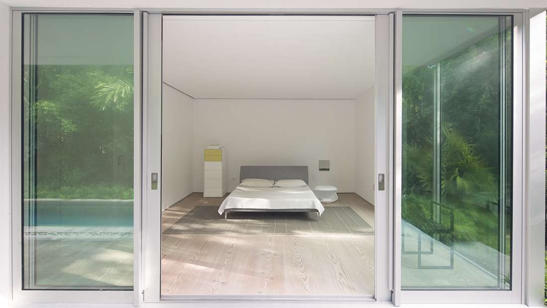 Minimalist bedroom with large sliding glass doors opening to a private pool and lush greenery, featuring light wood flooring, low-profile bed with gray headboard, and simple white walls.