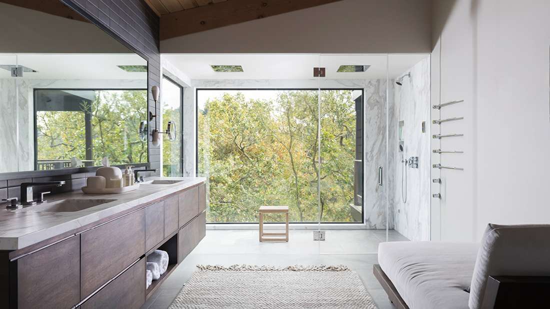 Luxury bathroom with double-sink vanity featuring dark wood cabinetry and marble countertop, large wall mirror, glass-enclosed shower with rainfall fixtures, and floor-to-ceiling windows overlooking lush green trees.