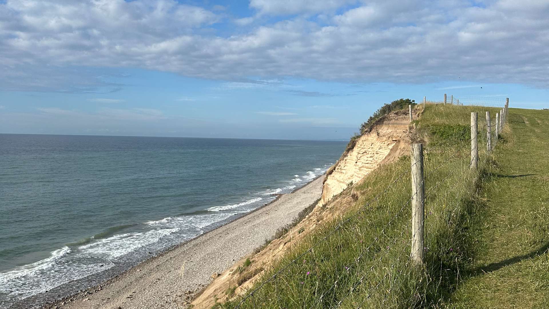Grassy cliff with wooden posts overlooking a rocky beach and calm ocean under a partly cloudy blue sky.