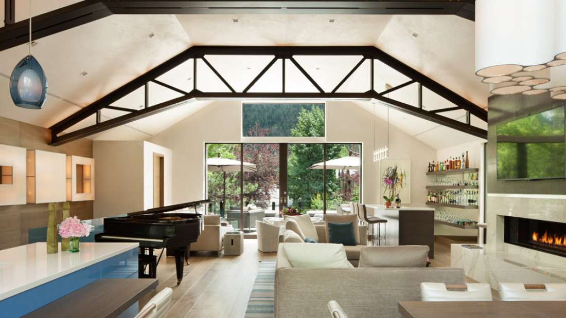 Spacious modern living area with vaulted ceiling and exposed beams, grand piano, sleek fireplace, and large glass doors opening to a lush outdoor patio.