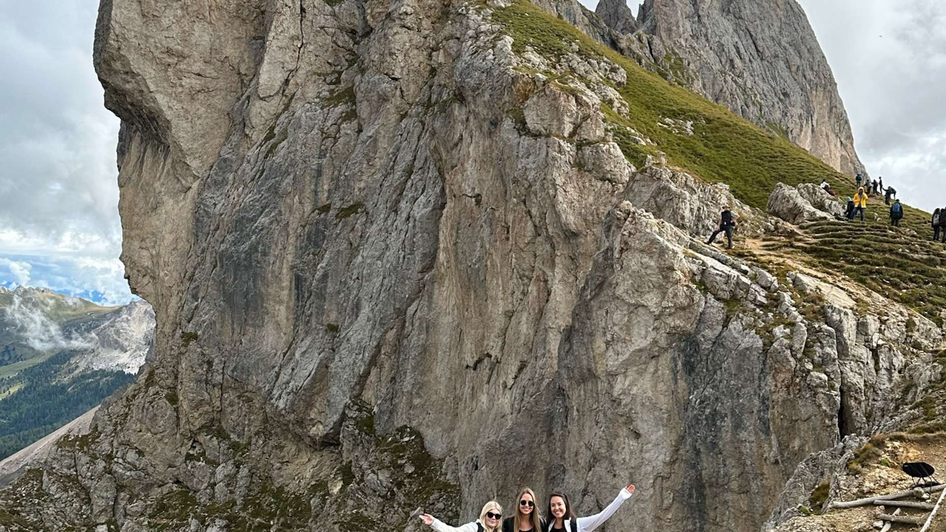 Three hikers standing on a rocky trail with arms raised in front of dramatic jagged mountain peaks at Seceda in the Dolomites under cloudy skies.
