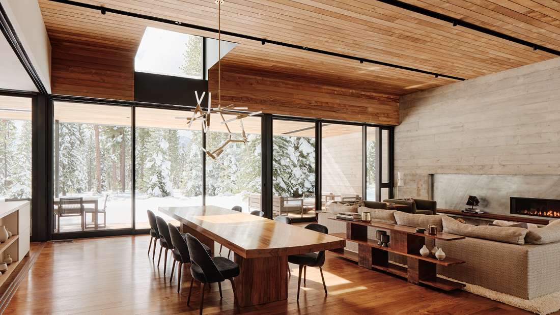 Luxury open-concept dining and living area with floor-to-ceiling glass walls, exposed wood ceiling panels, natural wood dining table with black chairs, and modern fireplace overlooking snowy forest views.