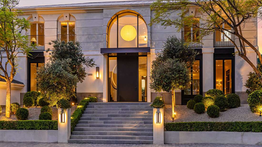 Luxury mansion exterior with grand arched glass entryway, symmetrical facade featuring tall windows and balconies, wide stone staircase with modern lanterns, and landscaped garden with trimmed hedges and accent lighting at dusk.