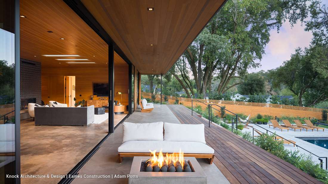 Luxury modern home with open-concept interior featuring wood-paneled ceiling, floor-to-ceiling glass walls, and outdoor terrace with fire pit lounge overlooking a swimming pool and landscaped garden.