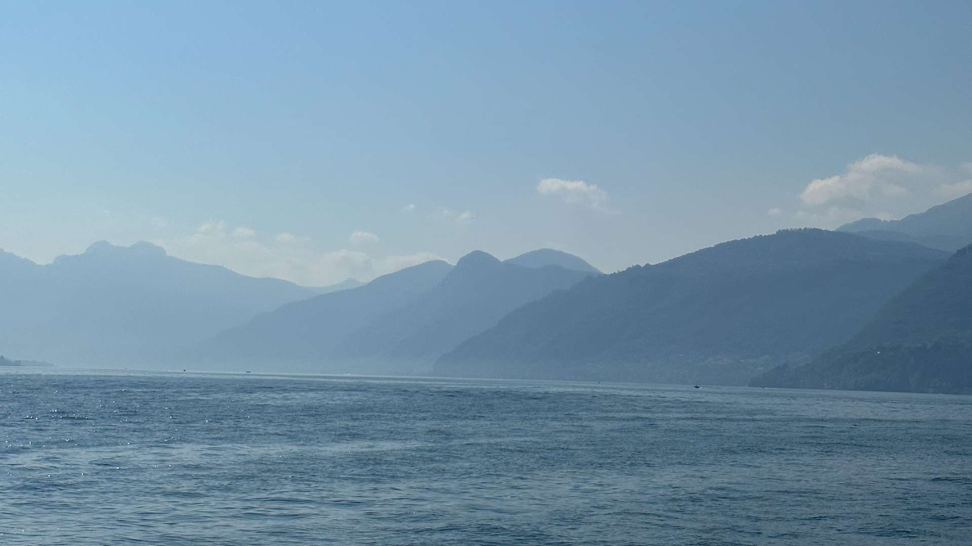 Calm blue lake with distant hazy mountain ranges under a clear sky with scattered clouds.