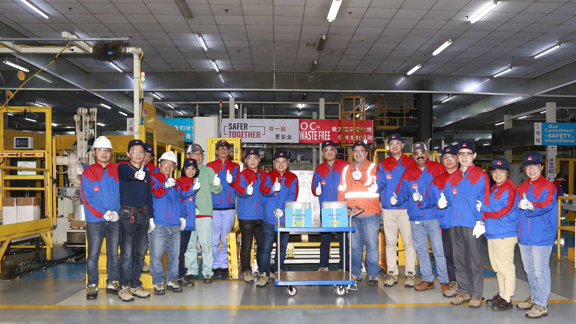 Factory workers in blue and red uniforms posing with safety signs and equipment inside an industrial facility.