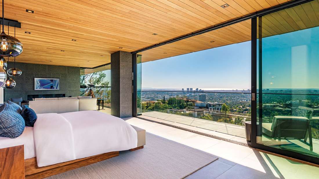 Luxury bedroom with floor-to-ceiling glass sliding doors opening to a balcony with panoramic city views, wood-paneled ceiling, modern pendant lights, and minimalist decor.