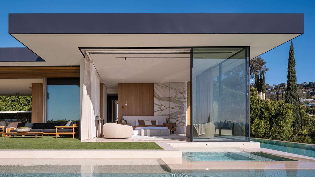 Luxury modern home with floor-to-ceiling glass walls, elegant bedroom interior, outdoor lounge chairs, and infinity pool at sunset.