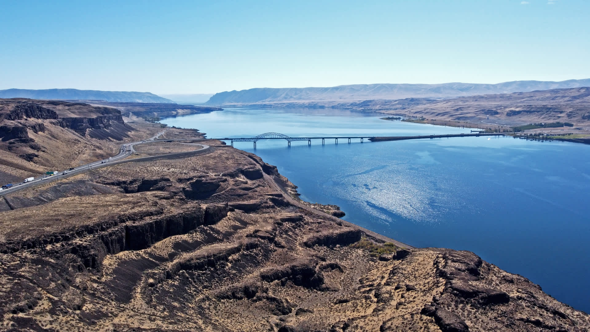   Aerial view of a wide river with a bridge crossing between rugged brown hills under a clear blue sky.