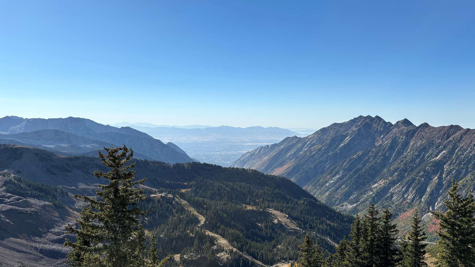 Expansive mountain landscape with steep rocky peaks, dense evergreen forests, and winding trails under a clear blue sky.
