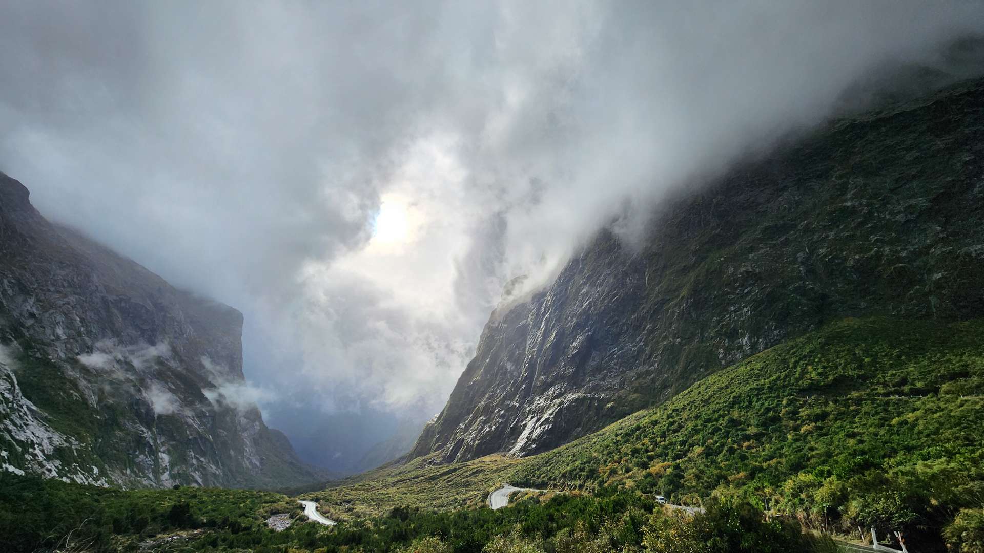   Winding mountain road through a lush green valley with towering cliffs and dramatic low-hanging clouds.
