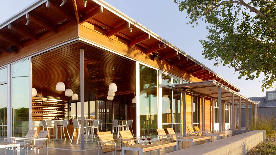 Modern glass-walled building with exposed wooden beams, sloped roof, and outdoor patio featuring wooden chairs and tables, illuminated by pendant lights during sunset.