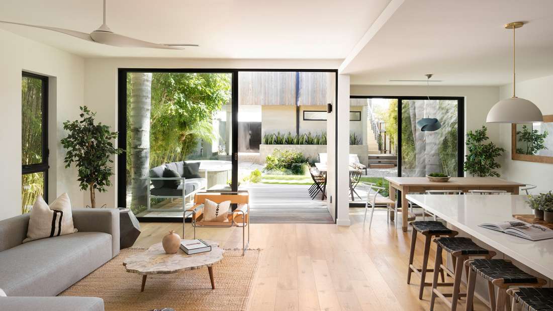 Bright open-concept living and dining area with modern furniture, light wood flooring, large sliding glass doors opening to a lush outdoor patio.